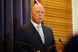 A bald man in a suit stands in front of an Australian flag, a blue curtain and a wooden-slatted window.