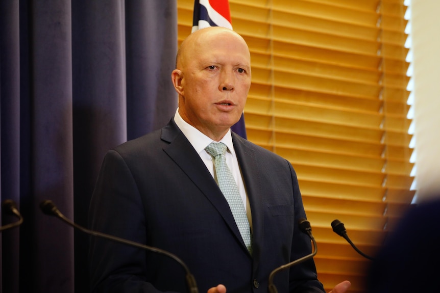 A bald man in a suit stands in front of an Australian flag, a blue curtain and a wooden-slatted window.