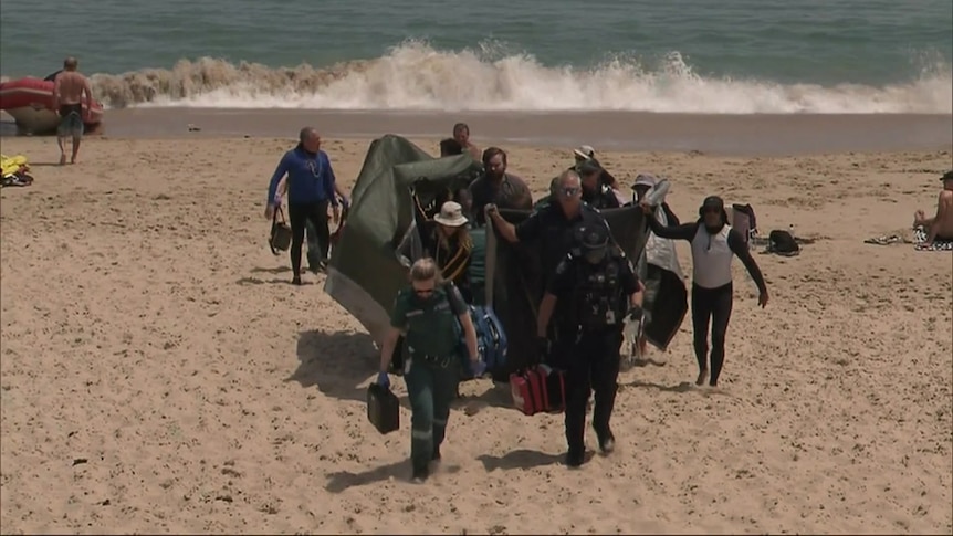 A team of paramedics carrying a woman along the beach away from the water.