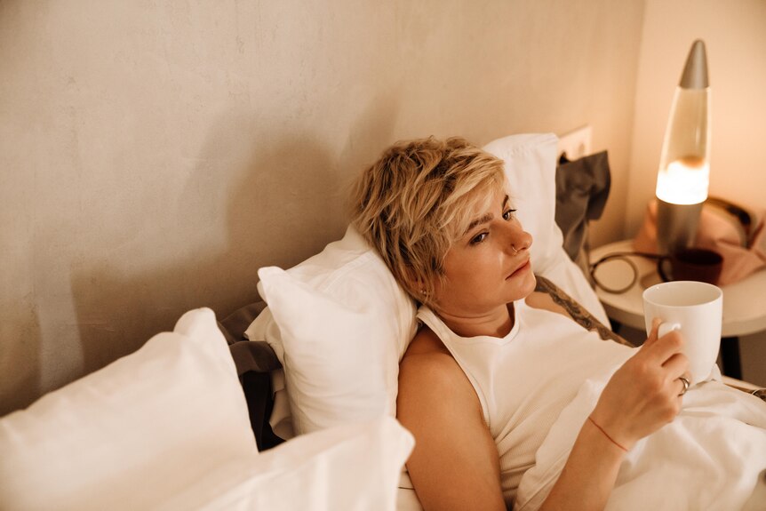 Woman with short blonde hair lying in bed looking unwell and holding a mug.