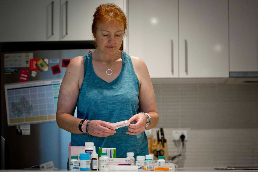 Janine Monty takes medication from a box while standing in her kitchen with all her medications on the counter in front of her.