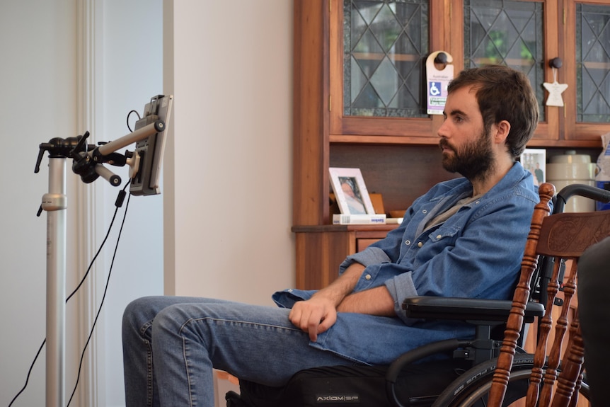 A man sits using eye gaze technology. He has brown hair, a beard and is wearing a denim shirt.