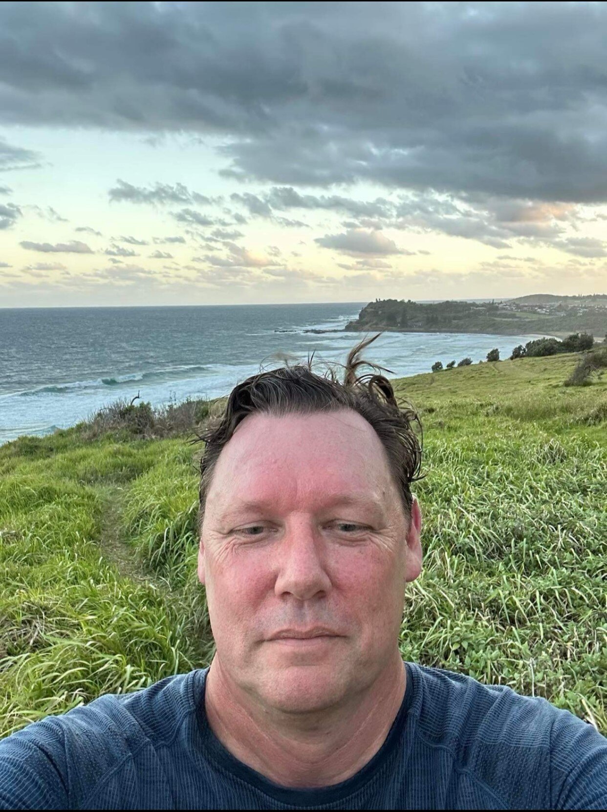 A man with brown hair takes a selfie with grass and open space behind him
