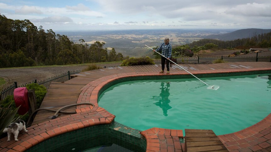 A woman skims a swimming pool in front of a sweeping vista of fields and hills.