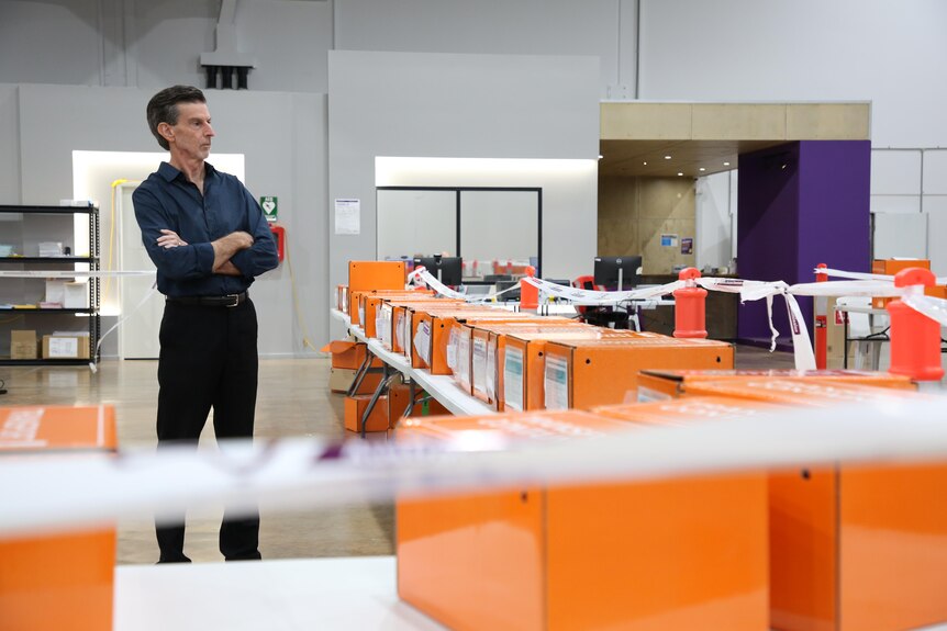 a man wearing a black business shirt and pants stands to the distance and looks at a collection of orange ballot boxes.