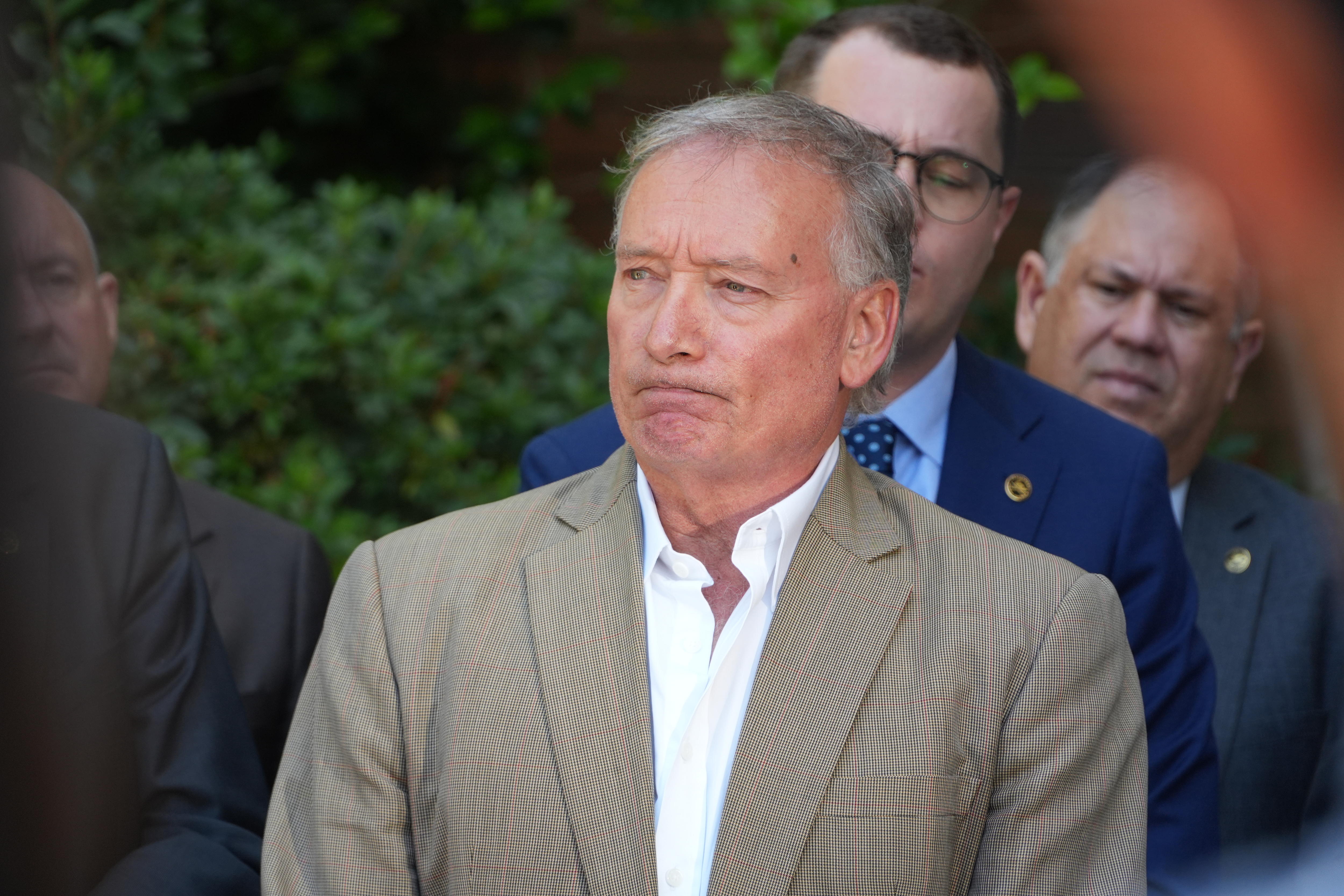 Un hombre de mediana edad, de pelo gris y vestido con un traje claro, se encuentra afuera, frente a otros hombres vestidos formalmente.