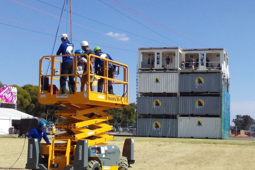 People with helmets are standing on a yellow lift, strapping a man into a harness and ropes. 