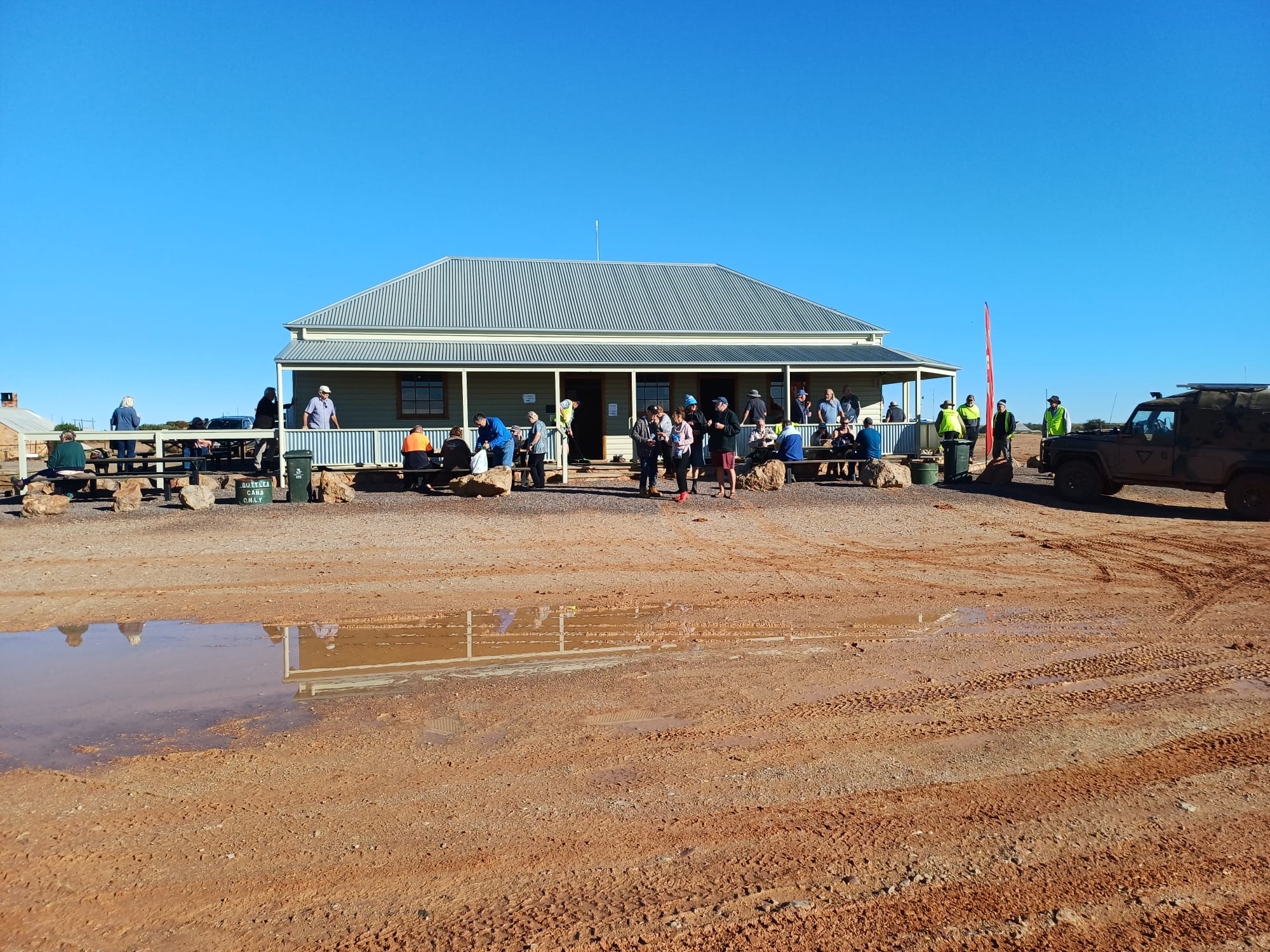 A weatherboard building with a verandah, with lots of people, under a blue sky and surrounded by red dirt.