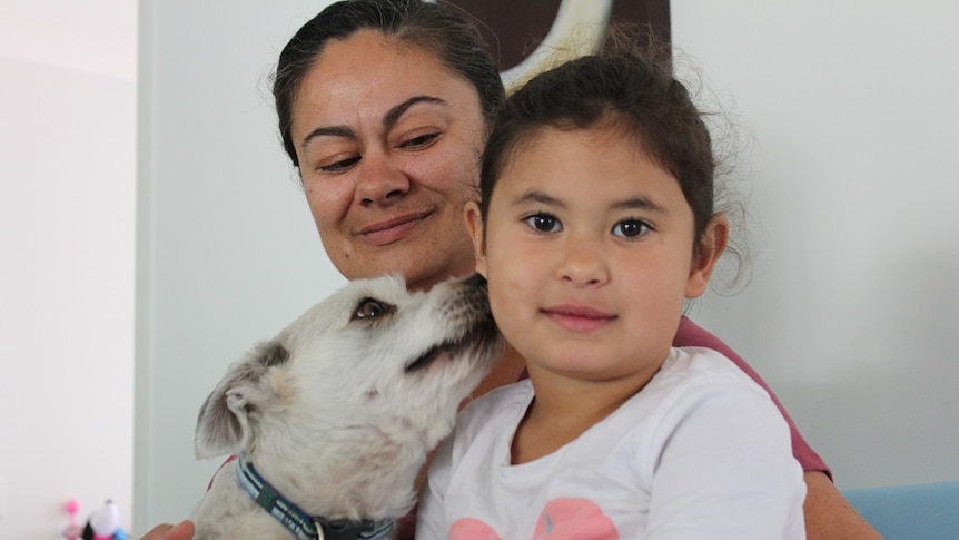 Mother, four-year-old daughter and dog sit closely together, with dog licking daughter's face.