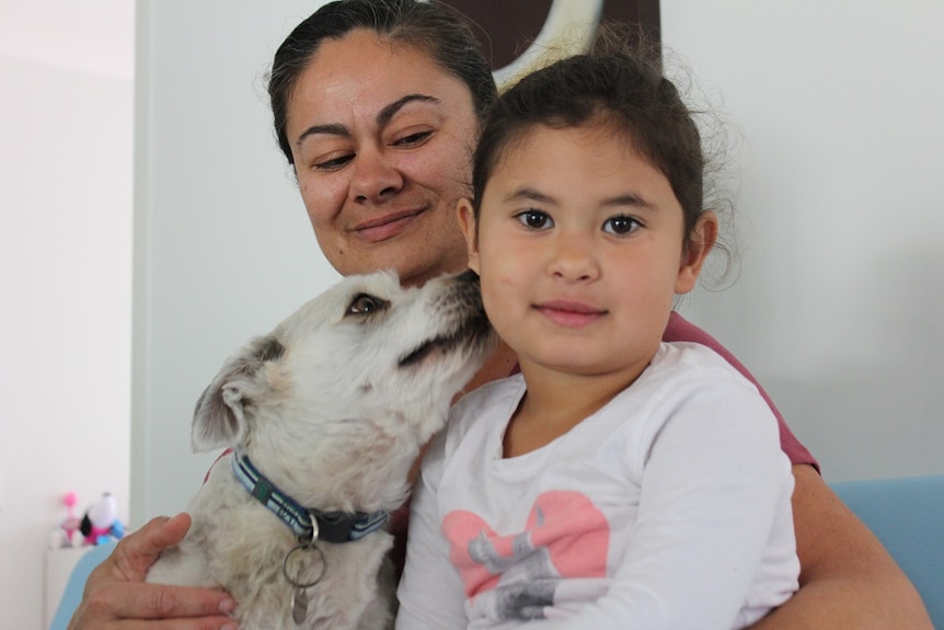 Mother, four-year-old daughter and dog sit closely together, with dog licking daughter's face.