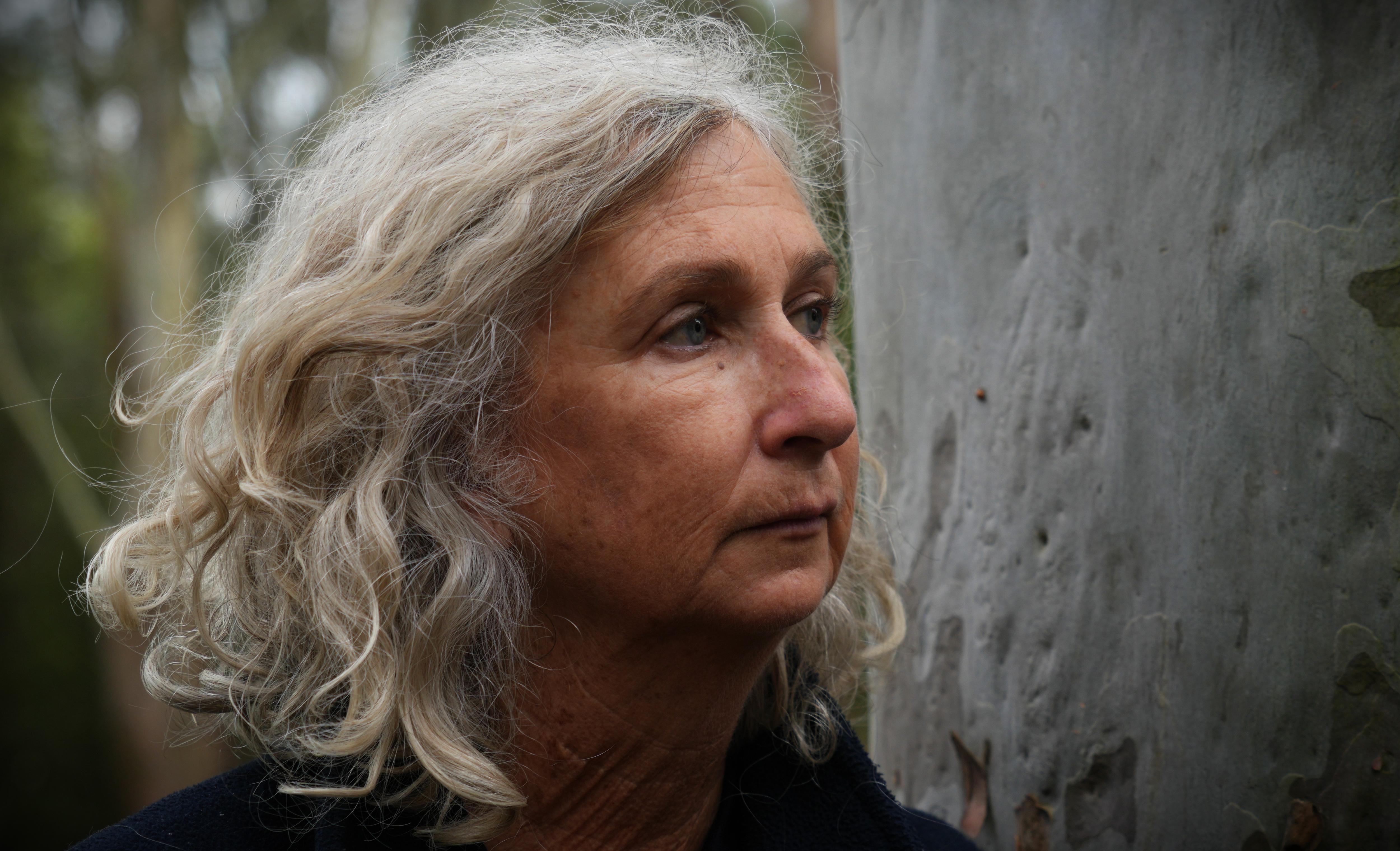Close up image of Kerry Walker with short hair looks off right of camera in front of tree trunk