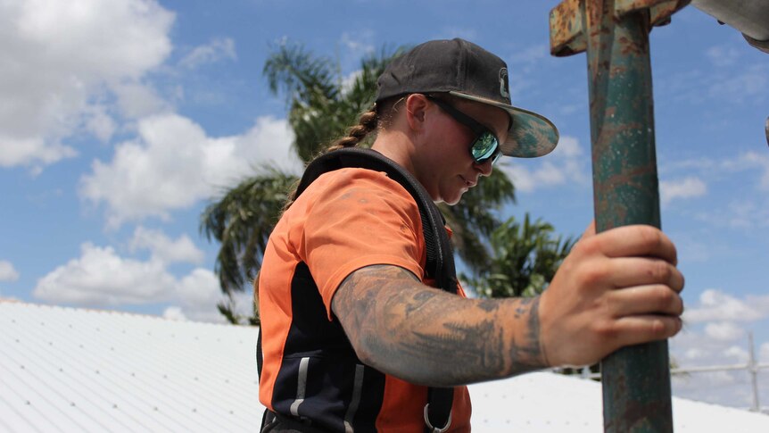 Carpentry apprentice Emily Bailey clutches scaffolding while on a roof, with blue skies and a palm tree behind her.