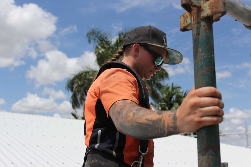 Carpentry apprentice Emily Bailey clutches scaffolding while on a roof, with blue skies and a palm tree behind her.