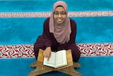 A woman sits on a prayer mat with an open Koran in front of her