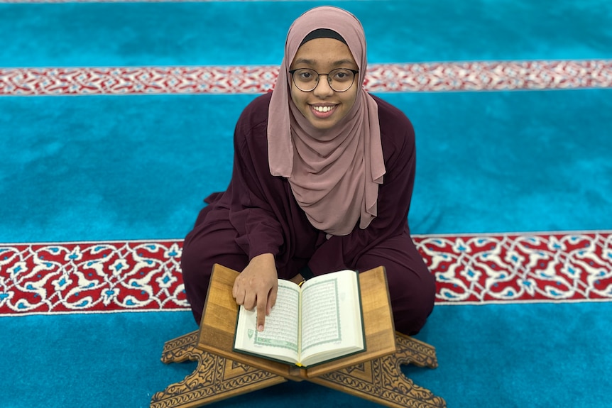 A woman sits on a prayer mat with an open Koran in front of her