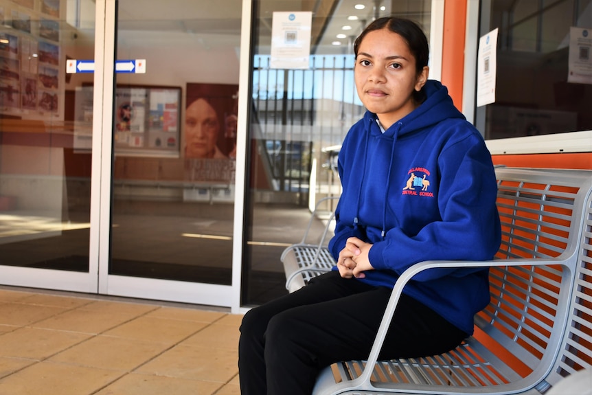 A teenage girl sits in a metal chair and looks to the camera.