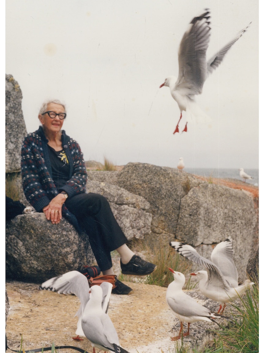 Edith Emery in her later life, sitting on a rock by the beach with seagullls in the foreground and one flying through the air