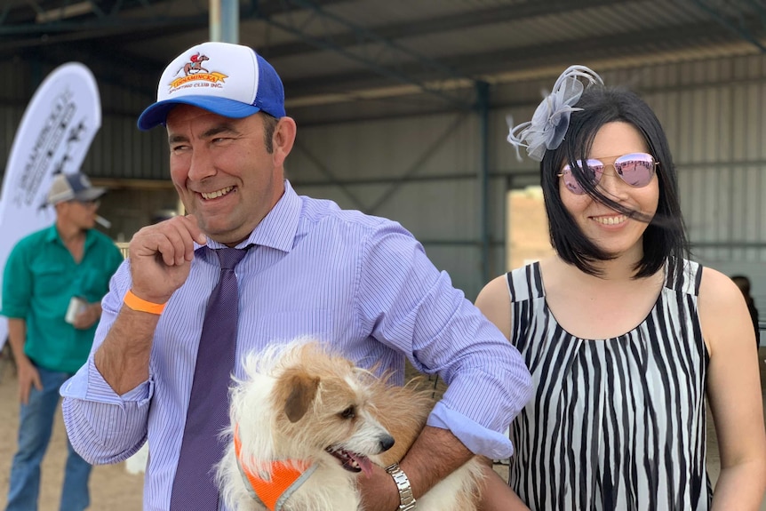 A man in a purple shirt and tie carries a small dog.  A woman with aviator sunglasses stands with him.