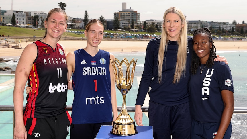 Four women's basketball players stand next to the World Cup trophy, with Bondi Beach behind them