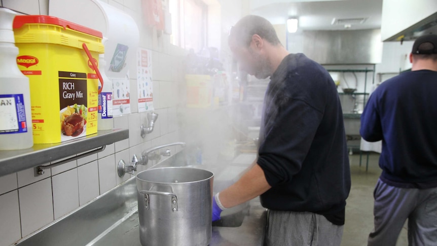 A man standing in a commercial kitchen with steam rising into his face, and a big pot to his left.