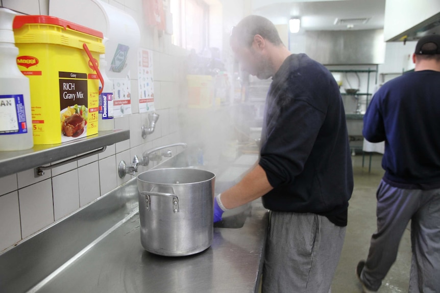 A man standing in a commercial kitchen with steam rising into his face, and a big pot to his left.
