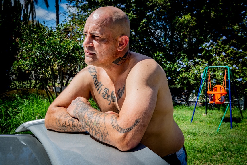Man leaning on a car, arms folded, shirtless. He was tattoos on his chest and arms.
