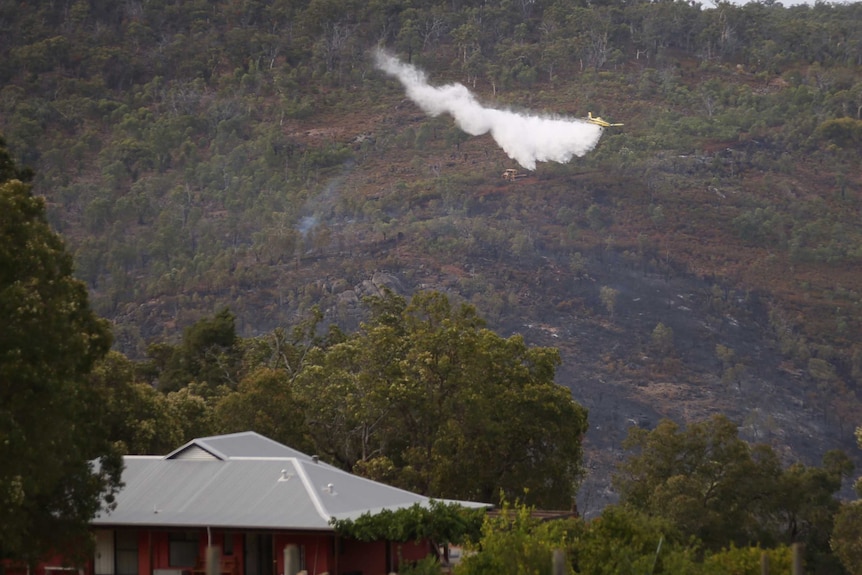 A water bombing plane drops water over an area of bushland with a house in the foreground.