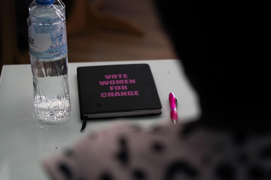 A black book with pink text VOTE WOMEN FOR CHANGE sits on a desk next to a bottle of water and pink pen