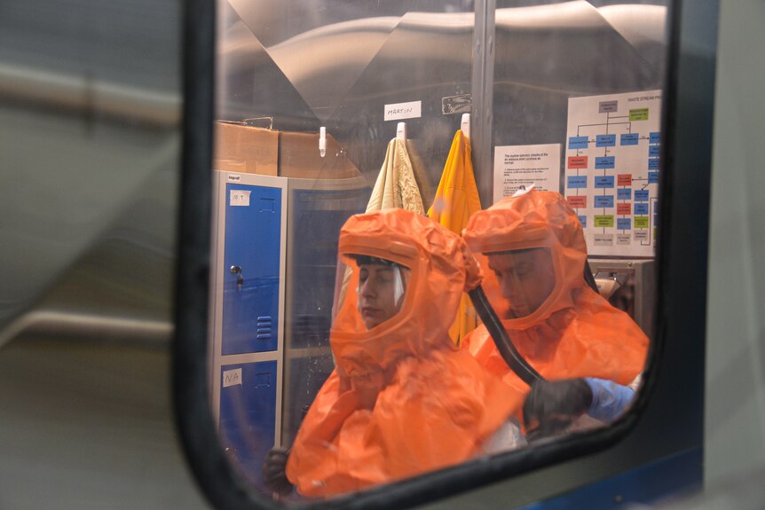 A male worker helps his female colleague put an orange hood and visor on, seen through a window