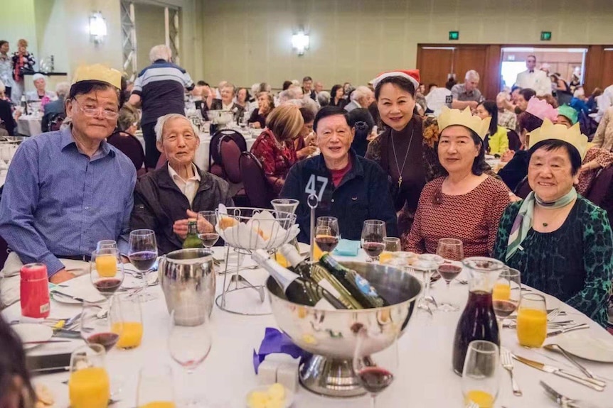 Three elderly men and three elderly women wear festive trinkets as they sit at a table with drinks, as part of a large gathering