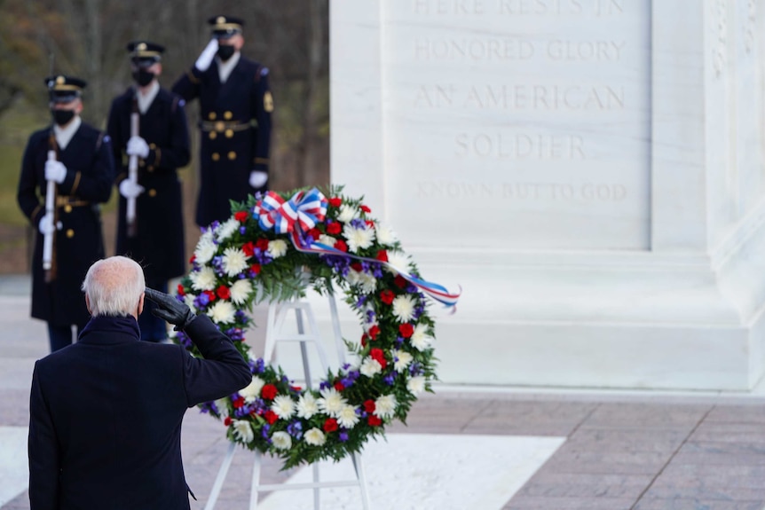 Back of Joe Biden head with right arm raised in salute in front of wreath.