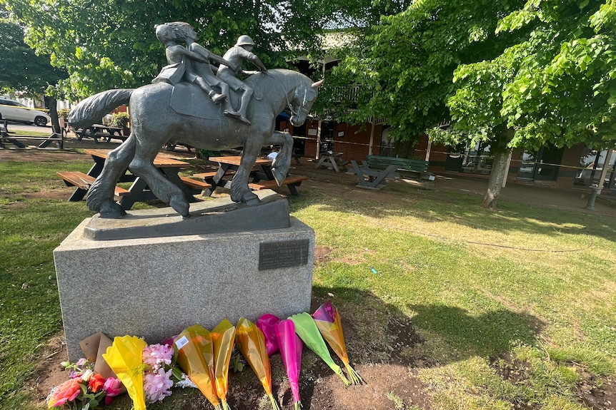 A statue in front of a grassed area with tables. Bouquets of flowers lie on the ground.