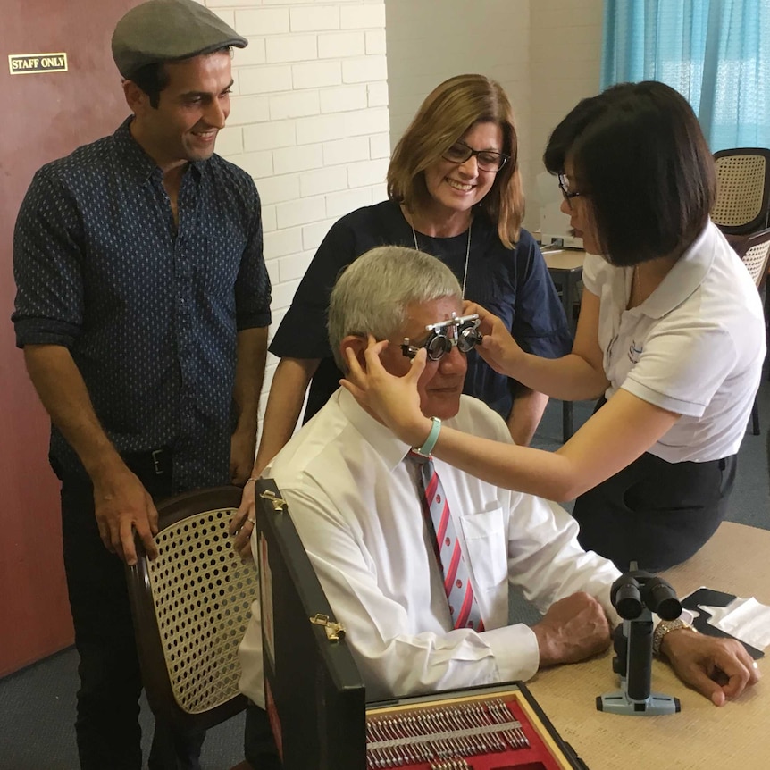 Ken Wyatt sits at a chair getting his eyes tested as two other people look on.