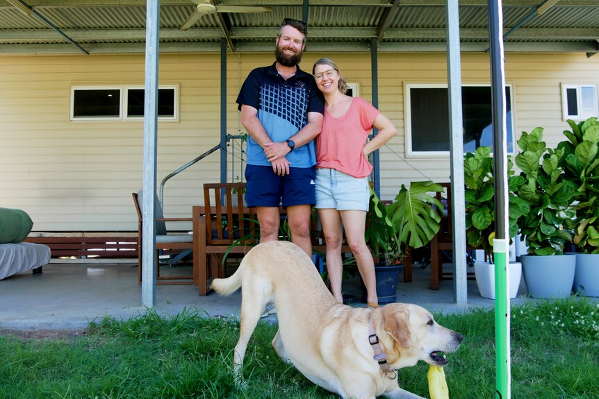 A man and woman smile in front of a house while their dog plays in front of them