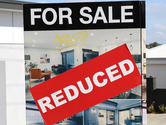 A 'for sale' sign with a red reduced sticker and two houses in the background