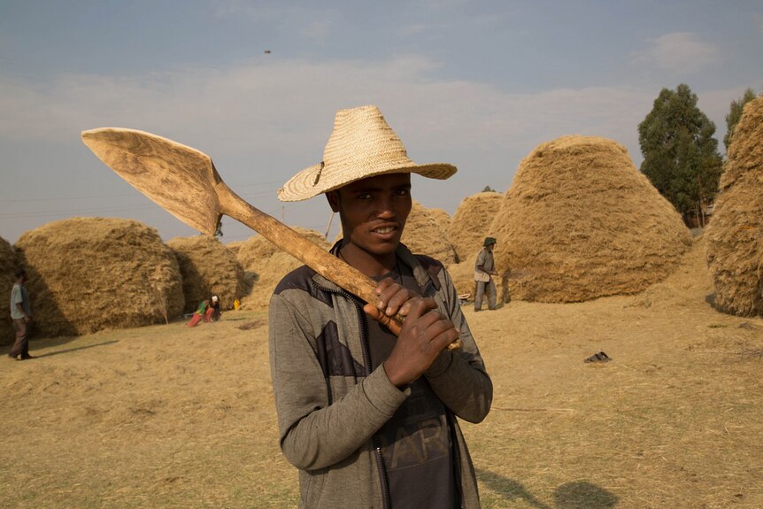 An Ethiopian man holds a wooden shovel and wears a straw hat in front of several hay mounds.