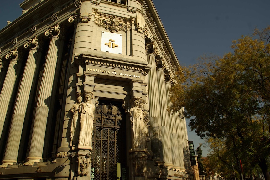 Looking up at a corner neoclassical building at dusk, which has two statues of women on either side of its wrought iron door.