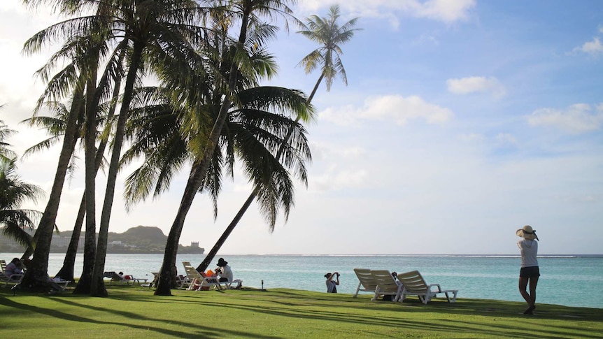 A beachfront scene in Guam, there is green grass, blue skies and palm trees. People are sitting around on deck chairs.