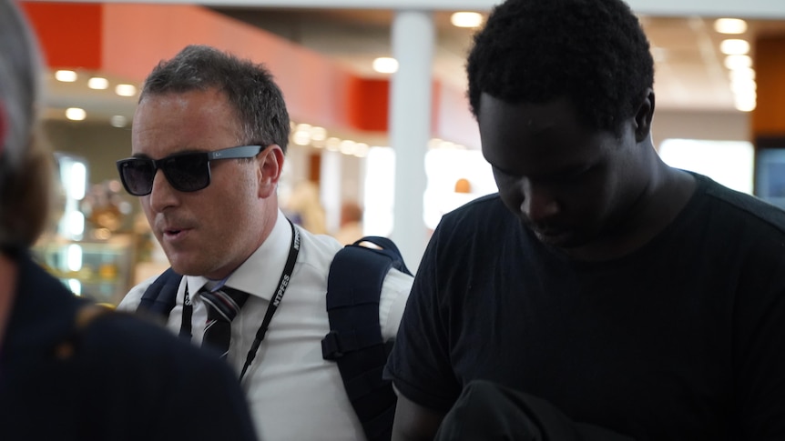 A young black man walks through the airport being ushered by a police officer in a white button up shirt