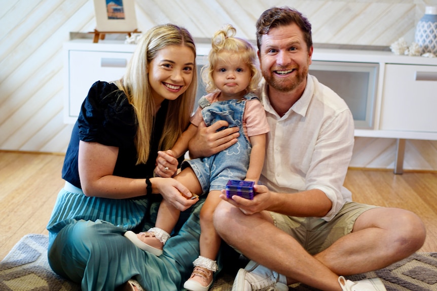 Lauren, Callum and their 18-month-daughter Elizabeth sit cross-legged on the floor, beaming at the camera.