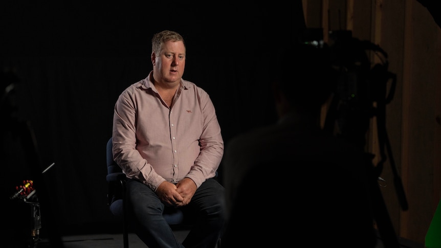 A man in a pink collared shirt sits in a dark room during an interview.