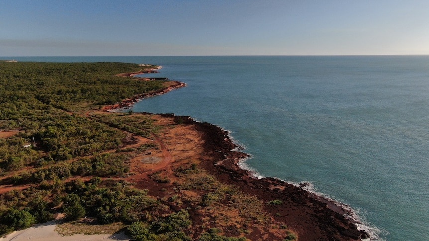 An aerial view of a strip of coastline, with red dirt and scrub meeting the ocean. 