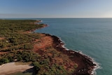 An aerial view of a strip of coastline, with red dirt and scrub meeting the ocean. 
