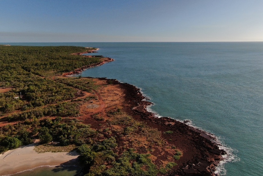 An aerial view of a strip of coastline, with red dirt and scrub meeting the ocean. 