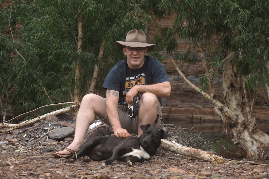 A man in a cowboy hat sits on a log with his dog lying down in front of him.