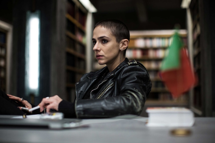 A scene from the movie The Translators with a woman in a leather jacket sitting at a desk.