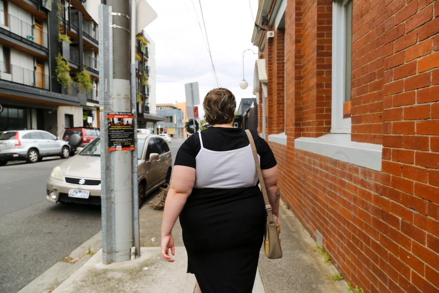 A woman walks down the street, facing away from the camera, wearing a backpack.