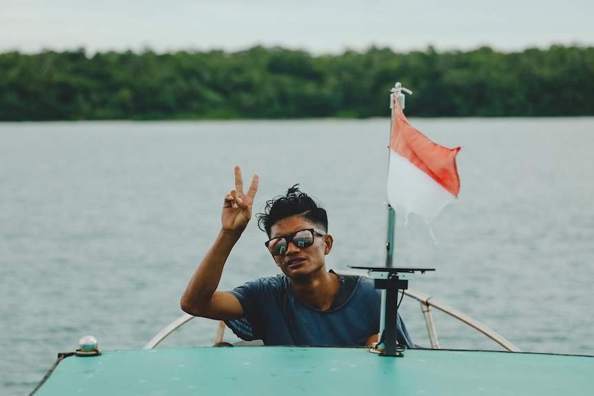 A man wearing a blue shirt and sunglasses gives the peace sign from the back of a boat with an Indonesian flag on it