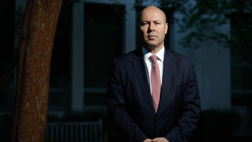 Josh Frydenberg stands in a suit and tie, looking at the camera with hands clasped in front. 