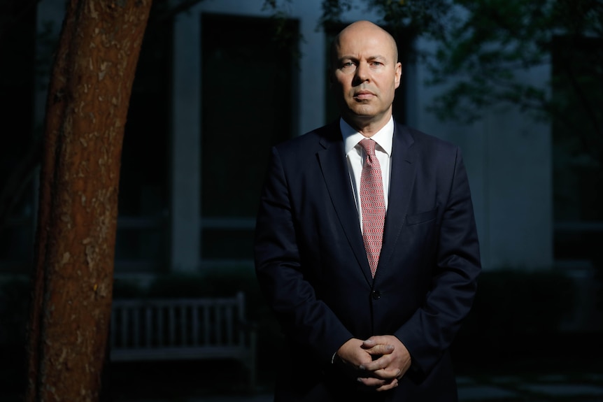 Josh Frydenberg stands in a suit and tie, looking at the camera with hands clasped in front. 